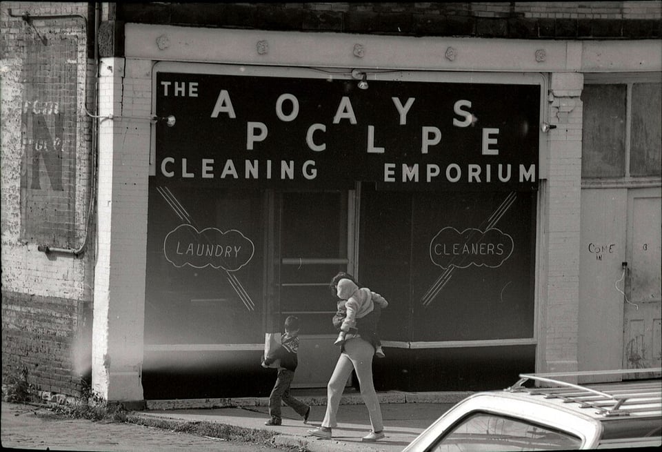 A black and white photo of mother and son walking in front of a store that says “The Apocalypse Cleaning Emporium” with neon lights in the windows that say “Laundry” and “Cleaners”.