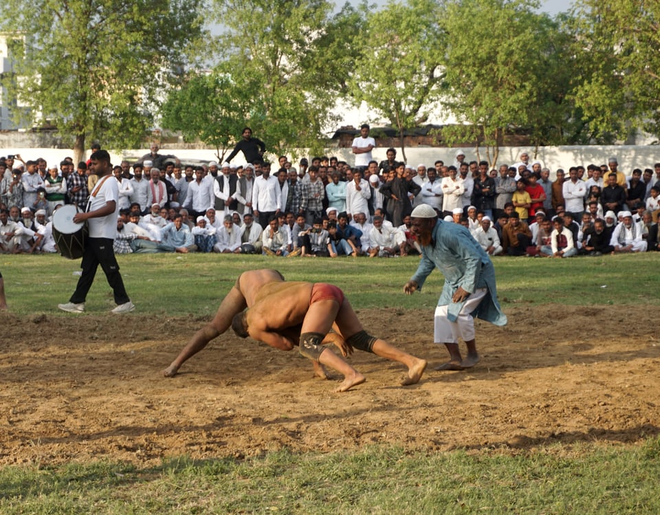 Two pehelwan(meaning champion) wrestling on a court made by clearing and smoothing earth on a flat ground. A referee in regular everyday traditional indian clothes watches closely nearby. A crowd anxiously watches in the background, a row standing on the ground and another two standing behind