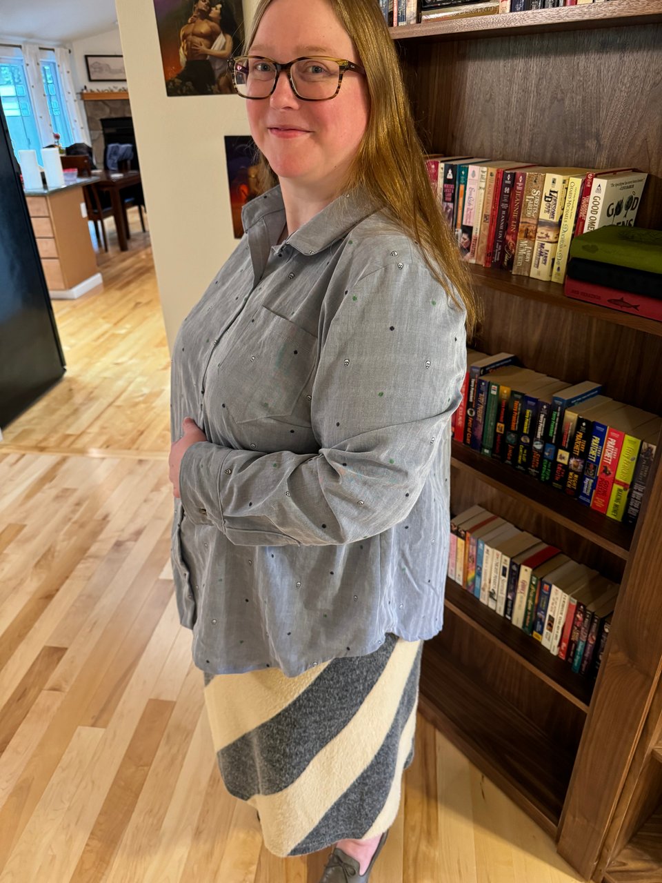 A white woman in a striped skirt stands in front of a bookcase — she’s wearing a soft blue button-up shirt dotted with tiny skulls and polka dots.
