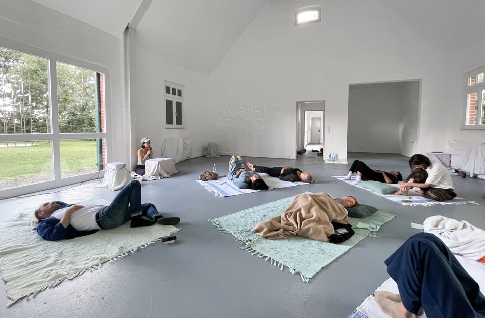 [People lying on blankets on the floor in a high-ceilinged, sunlit space. Charmaine sits in a corner reading into a microphone.]