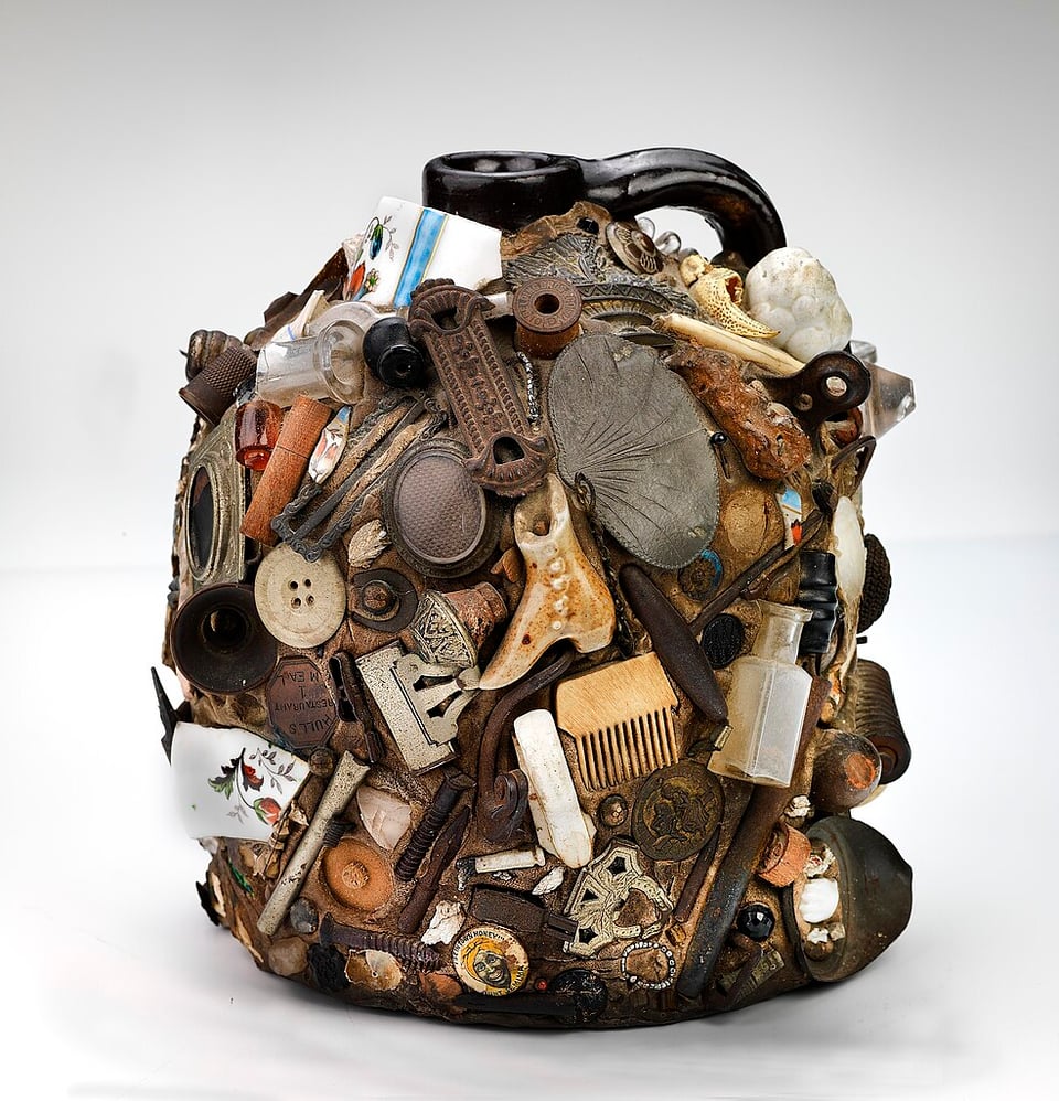 A jar coated in trinkets such as buttons, hair clips, screws, empty spools of thread. They are placed almost like a mosaic, with baubles covering all of it.