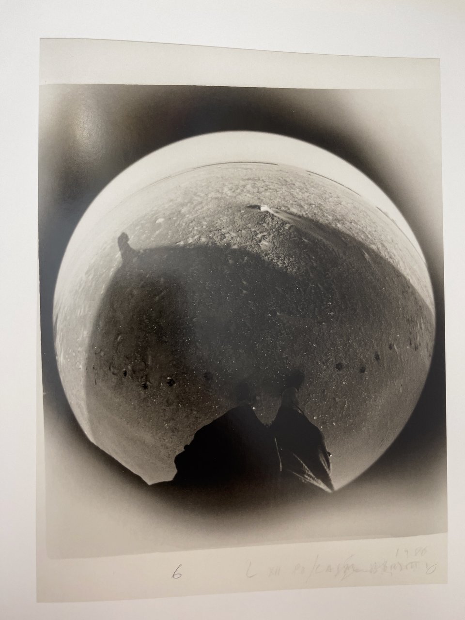 An image from Lunario, a photobook by Guido Guidi. The image is through a fisheye lens showing the photographers feet at the bottom and upside down. Above is a lunar like landscape. The image is black and white.