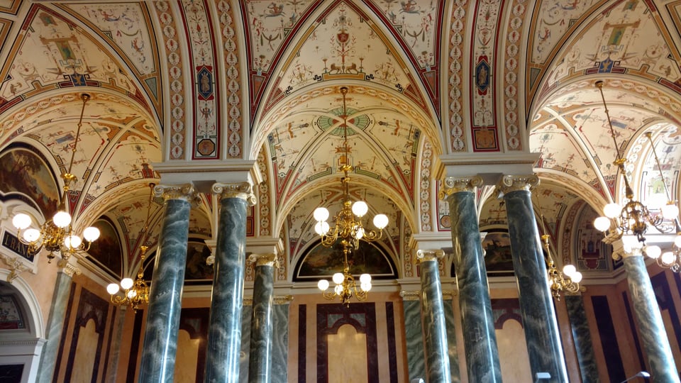 The interior foyer of the opera house. Green marble-look columns hold up the ribs of a vaulted ceiling. The ceiling is painted with designs that can't quite be made out at this resolution.