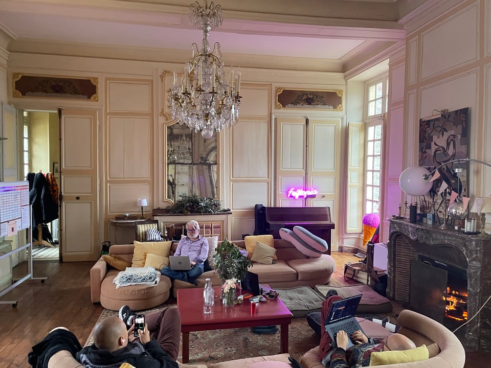 An ornate living room filled with sofas and pillows is occupied by three men using computers and phones.