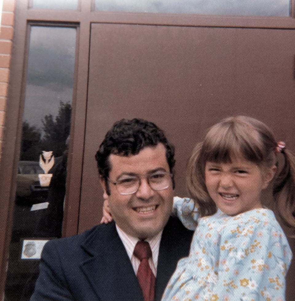 A white man with black hair and glasses is smiling and wearing a blue suitcoat with a striped shirt and red tie, holding a little girl who is also smiling, with brown hair put up in two pigtails and wearing a blue dress with little white and yellow flowers printed on it.