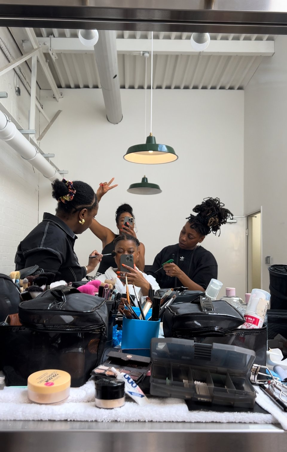 In a busy makeup room, I'm seated in the center, dressed in a white robe, receiving makeup from one artist while another styles my hair. A third artist behind me captures the moment on her smartphone. Around us, the table is cluttered with an array of makeup products and tools, reflecting a lively preparation scene.