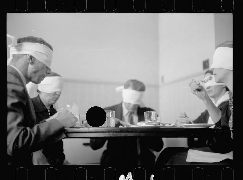 Five people sit around a table eating, wearing huge white blindfolds. There is a black circle in the image, made by a hole-puncher.