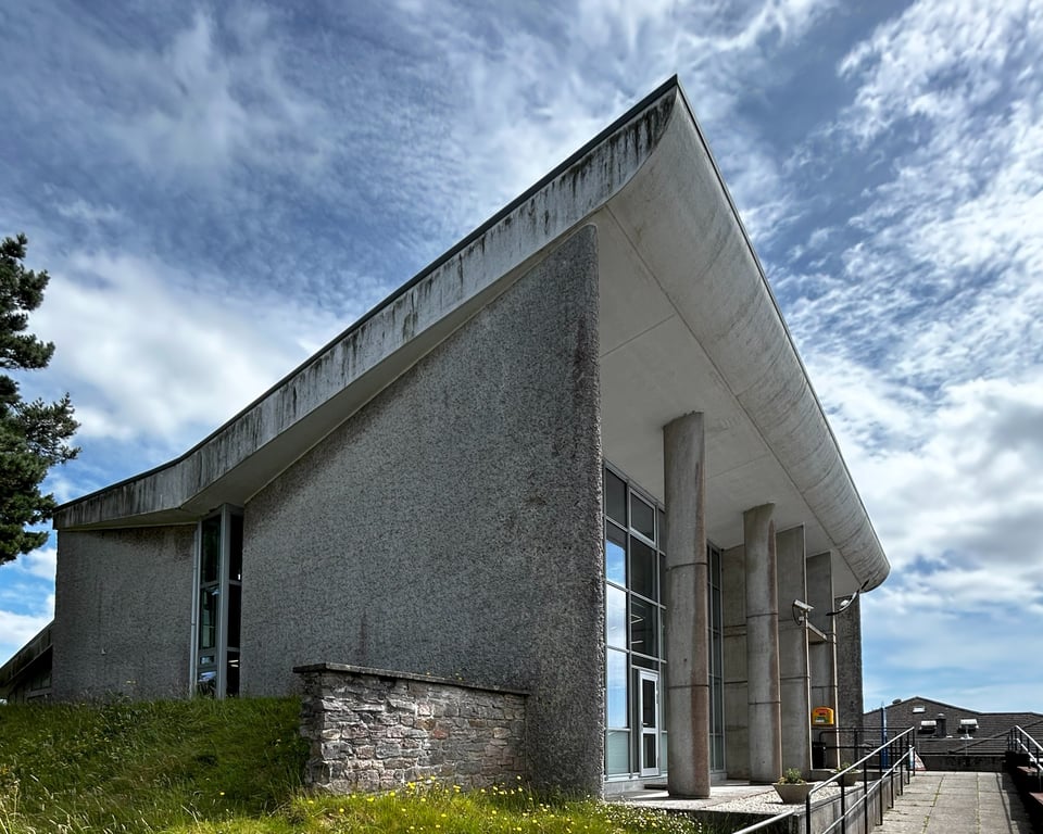 Concrete walls soar up to a double height butterfly roof. A curtain wall of glass is deeply recessed under the white swoop of the roof, and narrow oval pilotis hold it up.