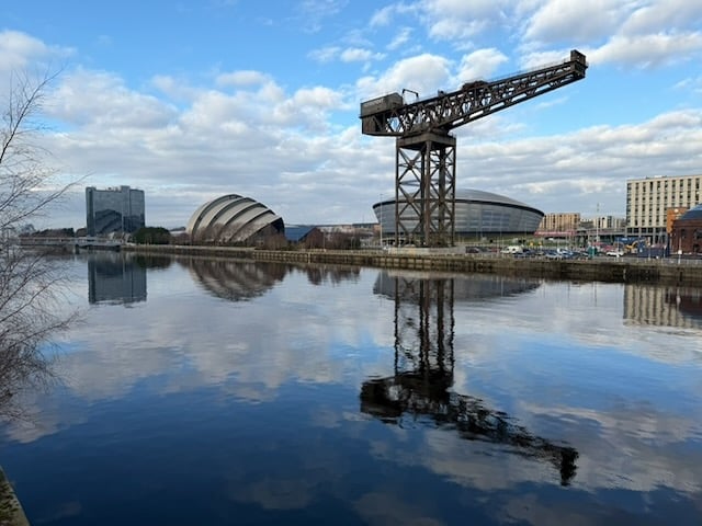 An image of the SEC Campus in Glasgow on a sunny day: three buildings and a crane, reflected in the still water of the River Clyde