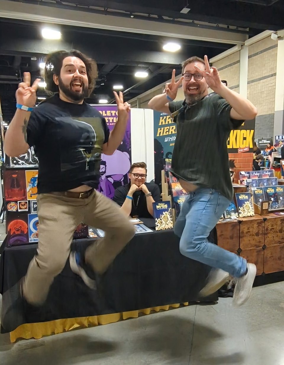 Two nerdy men jumping midair throwing up peace signs. Between them, a man holds his head up with his hands and a grin.