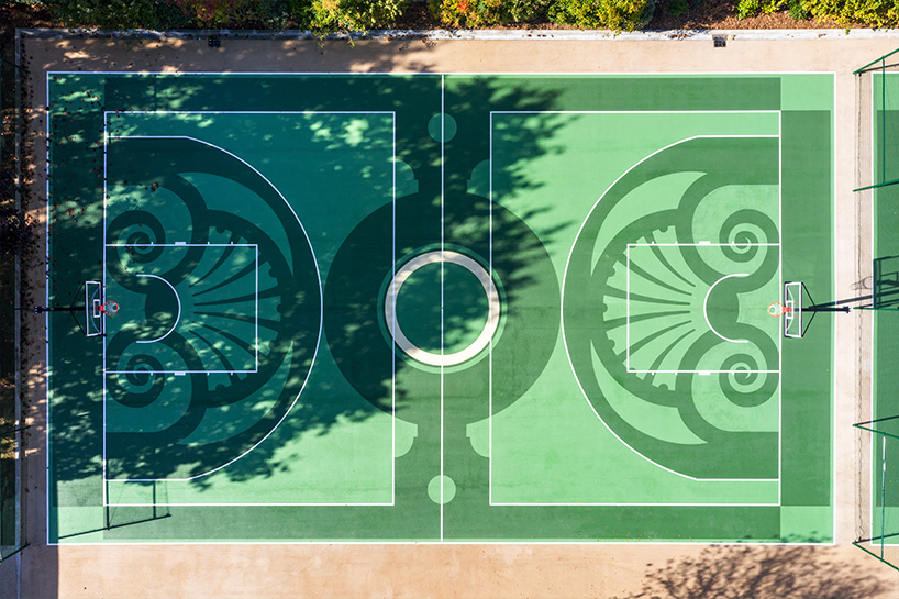 An aerial picture looking straight down on a basketball court. The basketball court is painted with a pattern of light green and dark green. There is a thick border of green around the edge of the court, and in the center a big dark circle. facing each net is a semi circle with an ornate pattern of curly cues, and what look like sheaves of thick wheat. It has the appearance of looking like a type of a garden. Around the edge of the court, off the cement, our planted trees.