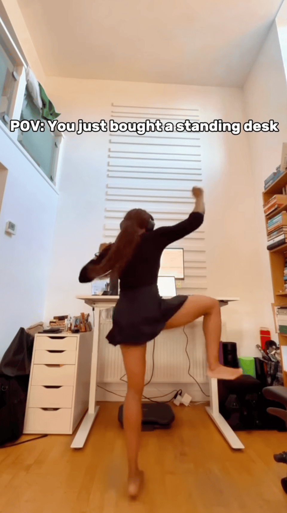 A woman dancing energetically in a home office in front of a standing desk, lifting one leg and swinging her arms, with text overlay reading “POV: You just bought a standing desk.” The space includes a white desk, drawers, bookshelves, and a laptop setup.