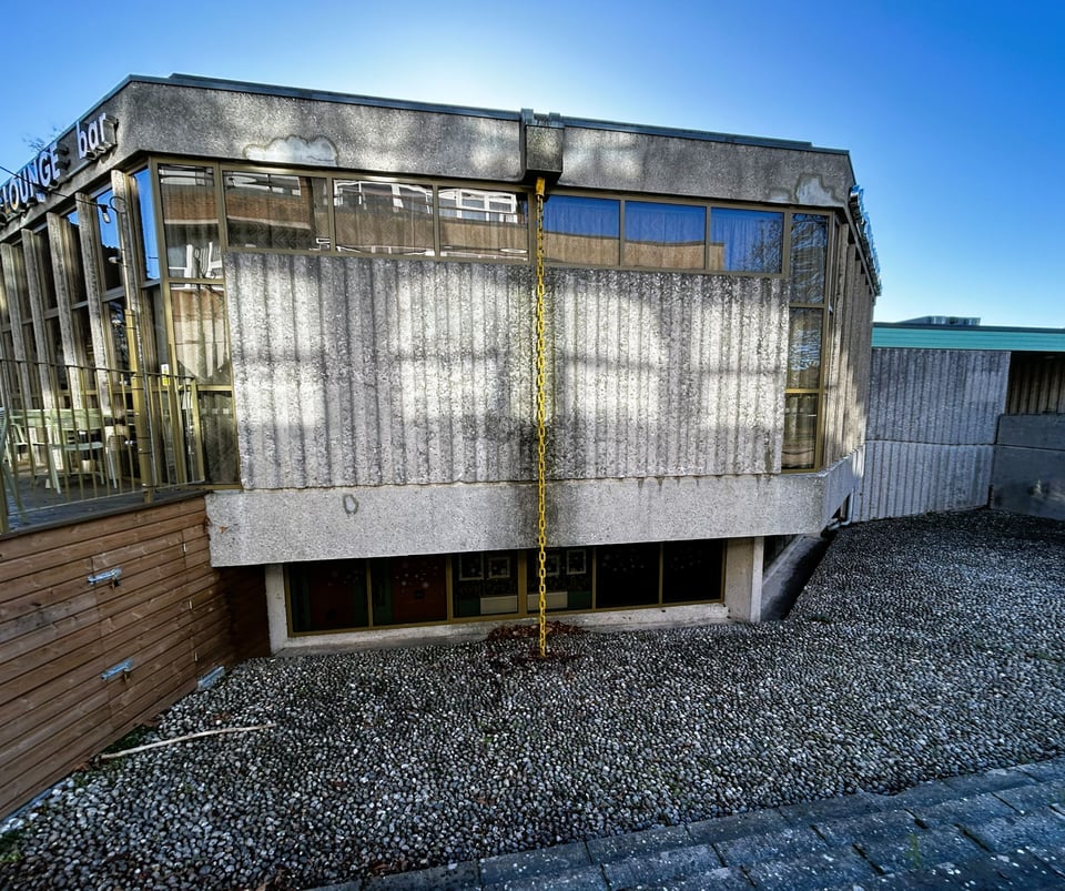 A ribbed concrete wall jets out over a lower ground floor strip window. The concrete panel has glass strip windows framing it. A chain runs from the gutters to the ground, and has been painted mustard yellow.