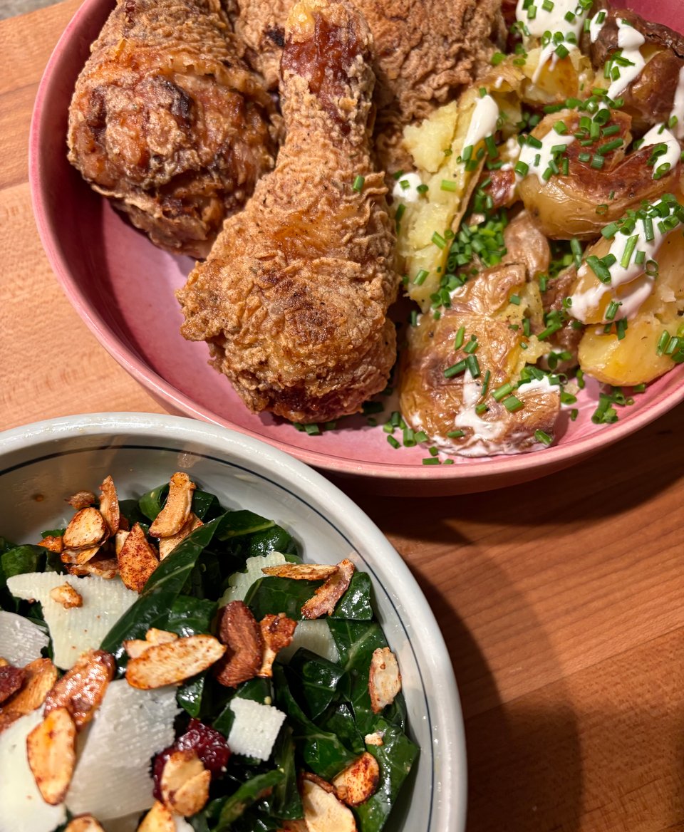 Lower corner depicts a light blue-gray bowl filled with a collard green salad topped with candied almond, dried cranberries, and shaved Parmesan. Top right contains a pink high-sided plate filled with crispy buttermilk fried chicken and a helping of small potatoes covered in salt-and-vinegar sauce and chives.