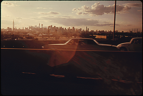 The skyline of New York City is behind a highway filled with cars.
