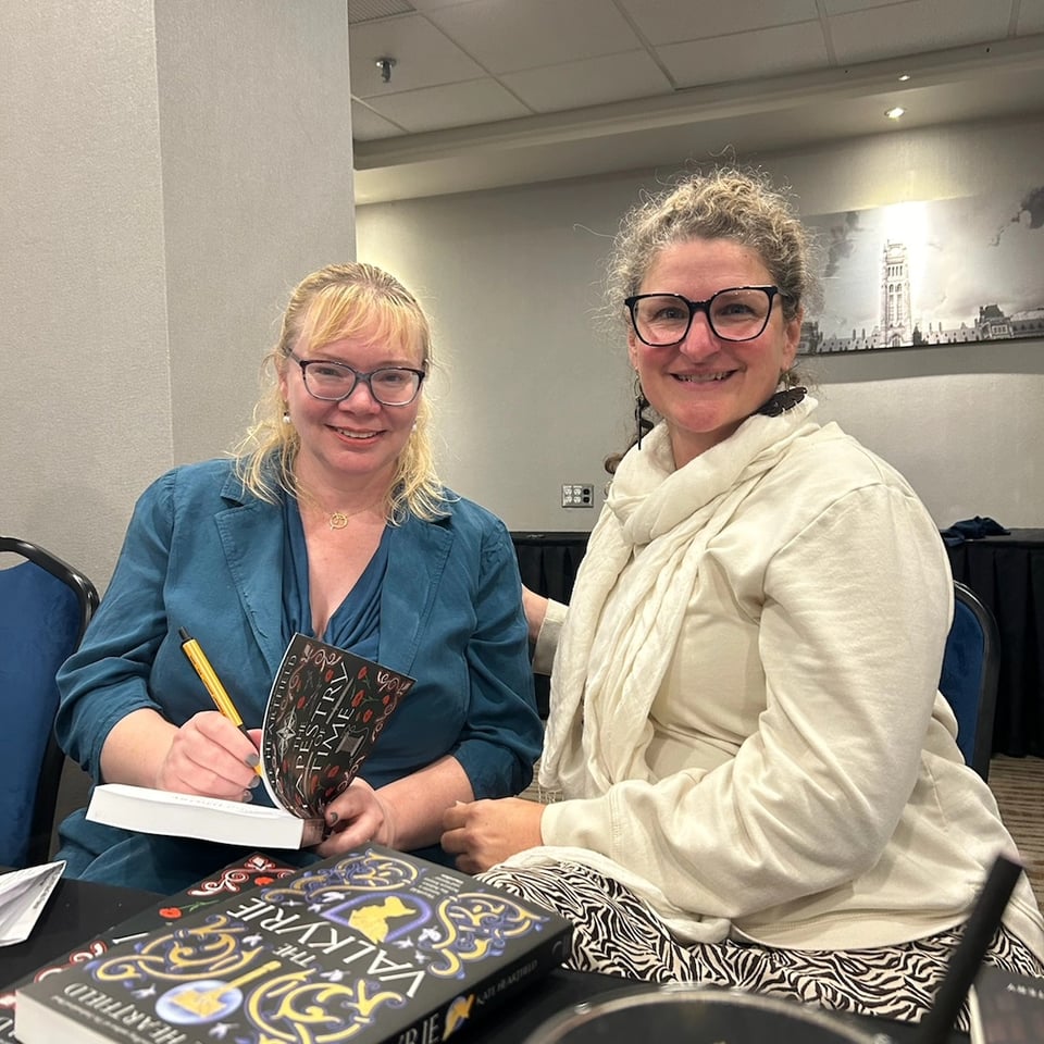 Kate in a blue suit signing a copy of The Tapestry of Time with Mika Weaver next to her.