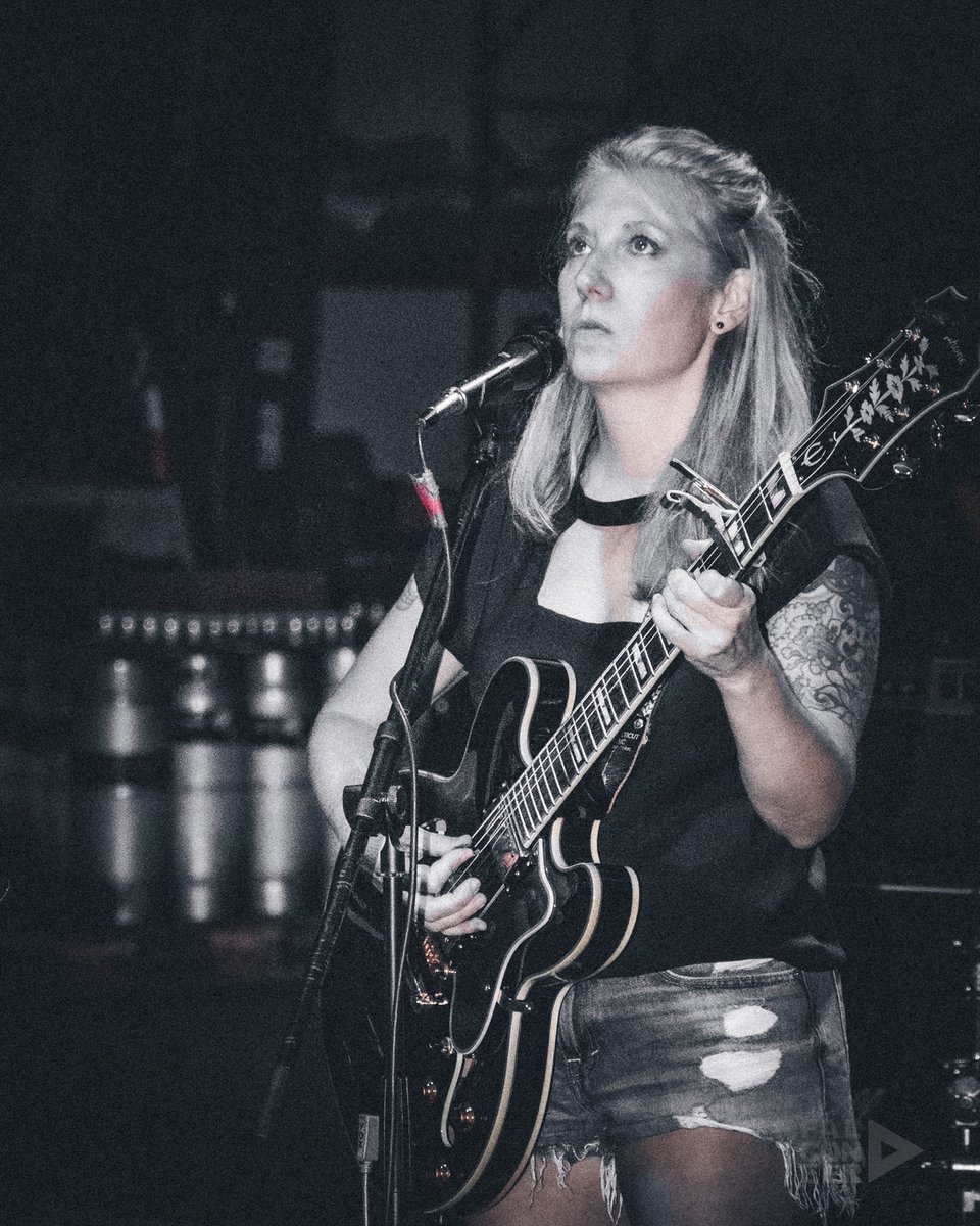 A woman with blond hair in a black tank top and cutoffs looks into the distance while she plays a black Epiphone semi-hollow body guitar
