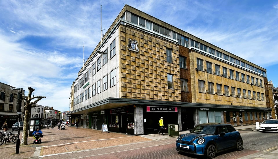 A large building taking up most of a block. Its front elevation is slabs of concrete or granite with square windows above a canopy. It's side elevation is more mixed, with a wall like a chequerboard made of bas relief squares, then a brick stairwell, then plain stone facade. At the top a run of ribbon windows is set back.