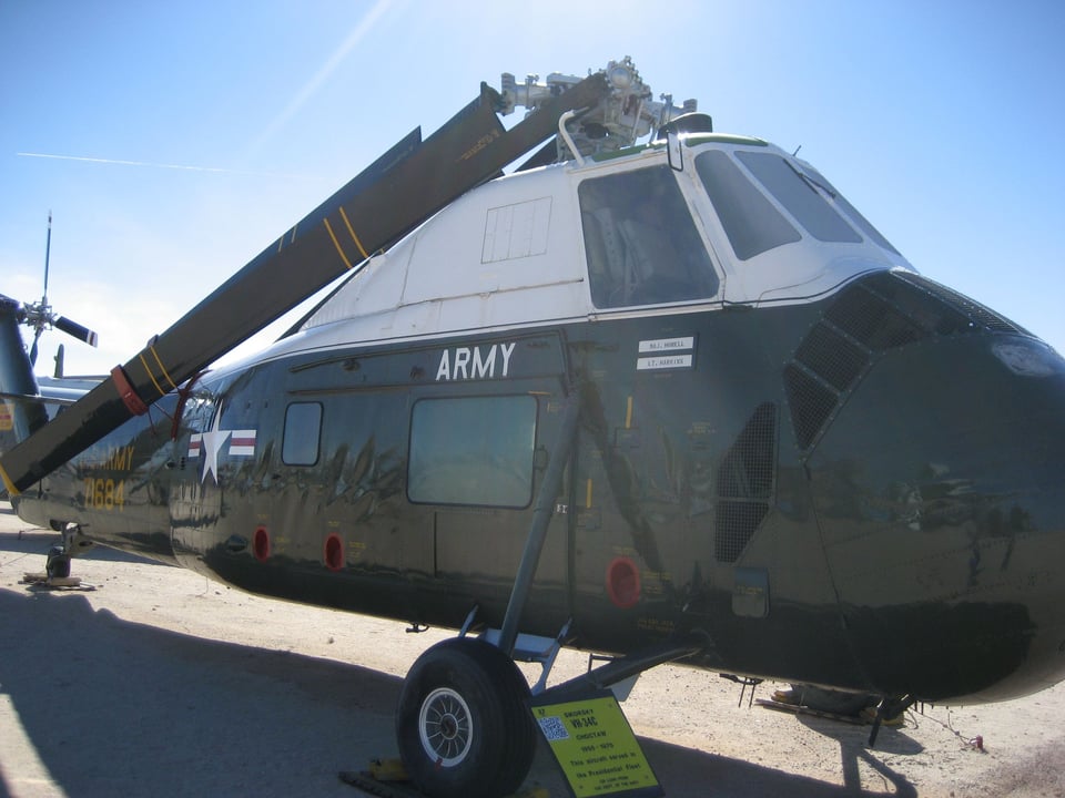 A helicopter painted in US Army colors, the rotors folded inwards, parked on light colored desert soil.