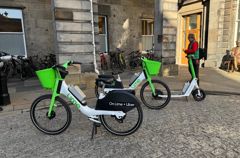 Two white and bright green hire ebikes from provider Lime sit in front of the city chambers in partial sunshine, atop grey setted ground. An electric scooter in the same colourway sits alongside; a councillor takes photographs of the assembled rides.