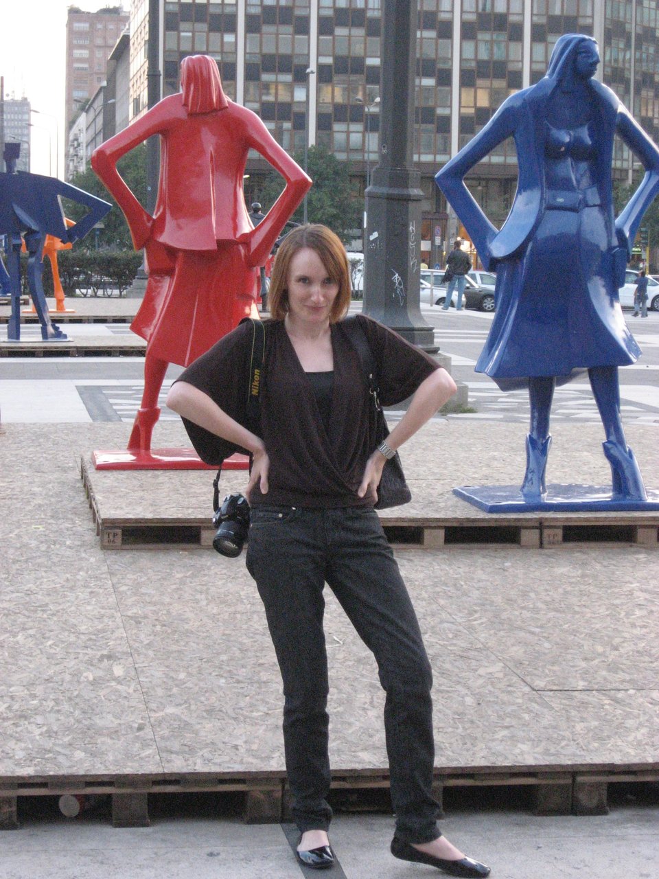 photo of beautiful white woman with red hair, standing with hands on hips and elbows out, mimicking the red and blue statues behind he