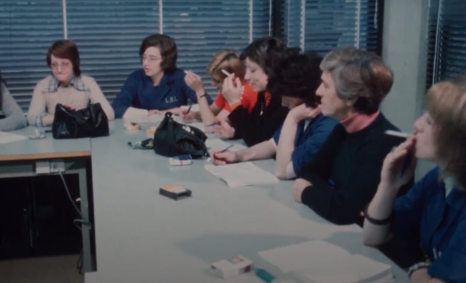 A photo of a meeting scene from the 60s or 70s, with seven women sitting around an office table layout, some of them smoking, some writing down notes, with windows in the background and closed blinds