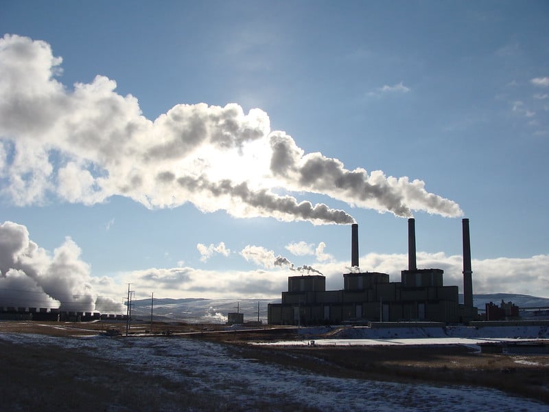 a three smokestack power plant with steam coming off the stacks and close by pools. The sun is filtered through one of the plumes of steam, putting the plant in silhouette. there are mountains in the background and a light amount of snow on the ground.