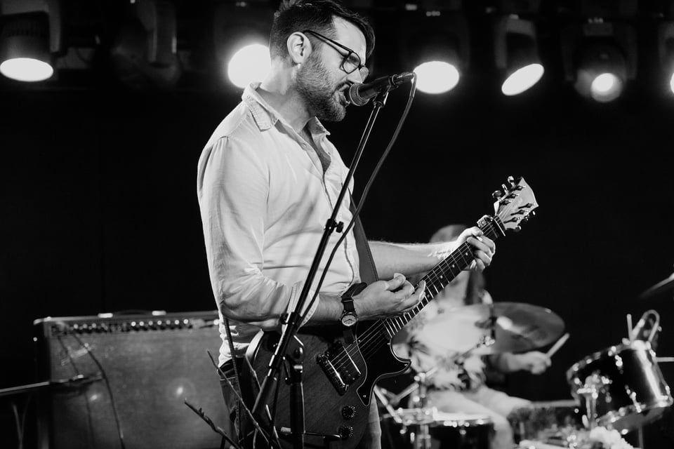 Steve is singing into the mic, mid-strum, shot from slightly below/to the side, in black and white