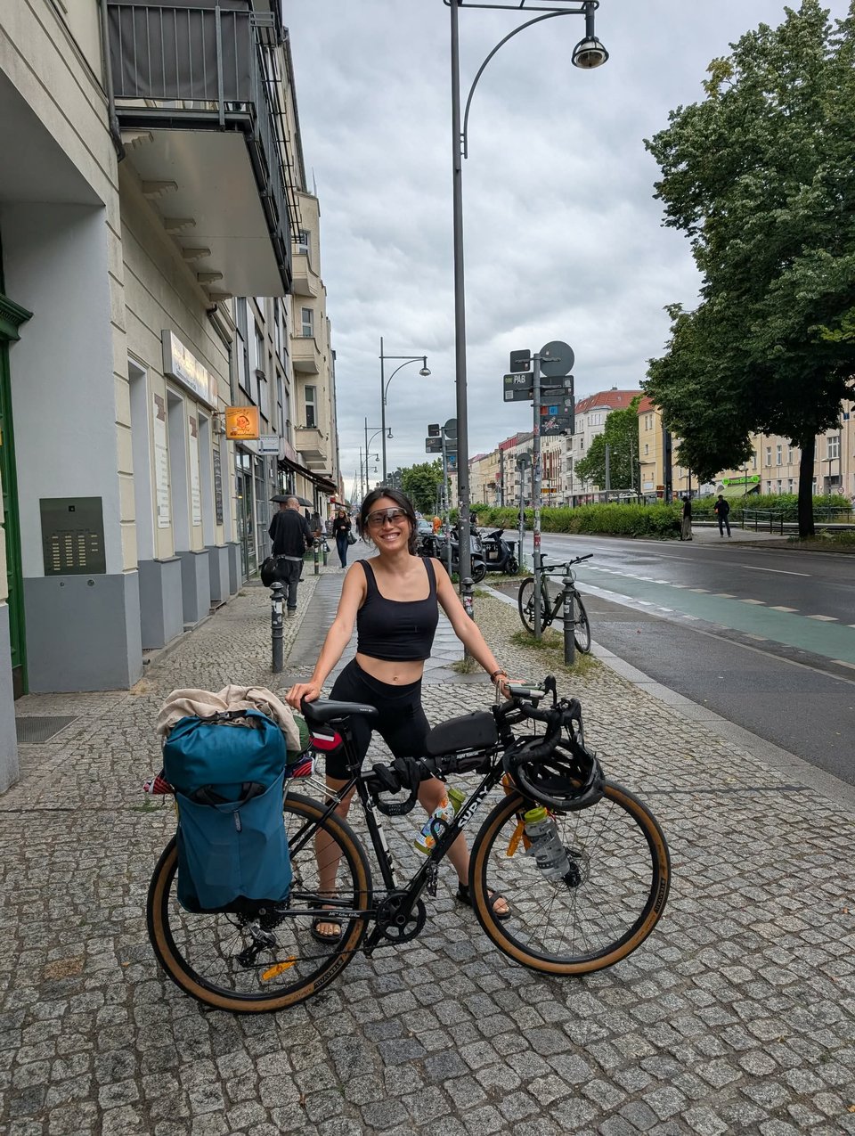 A woman standing on a cobblestone footpath with a bicycle laden with gear, wearing large cycling sunglasses, black cycling shorts, black crop top, and sandals.