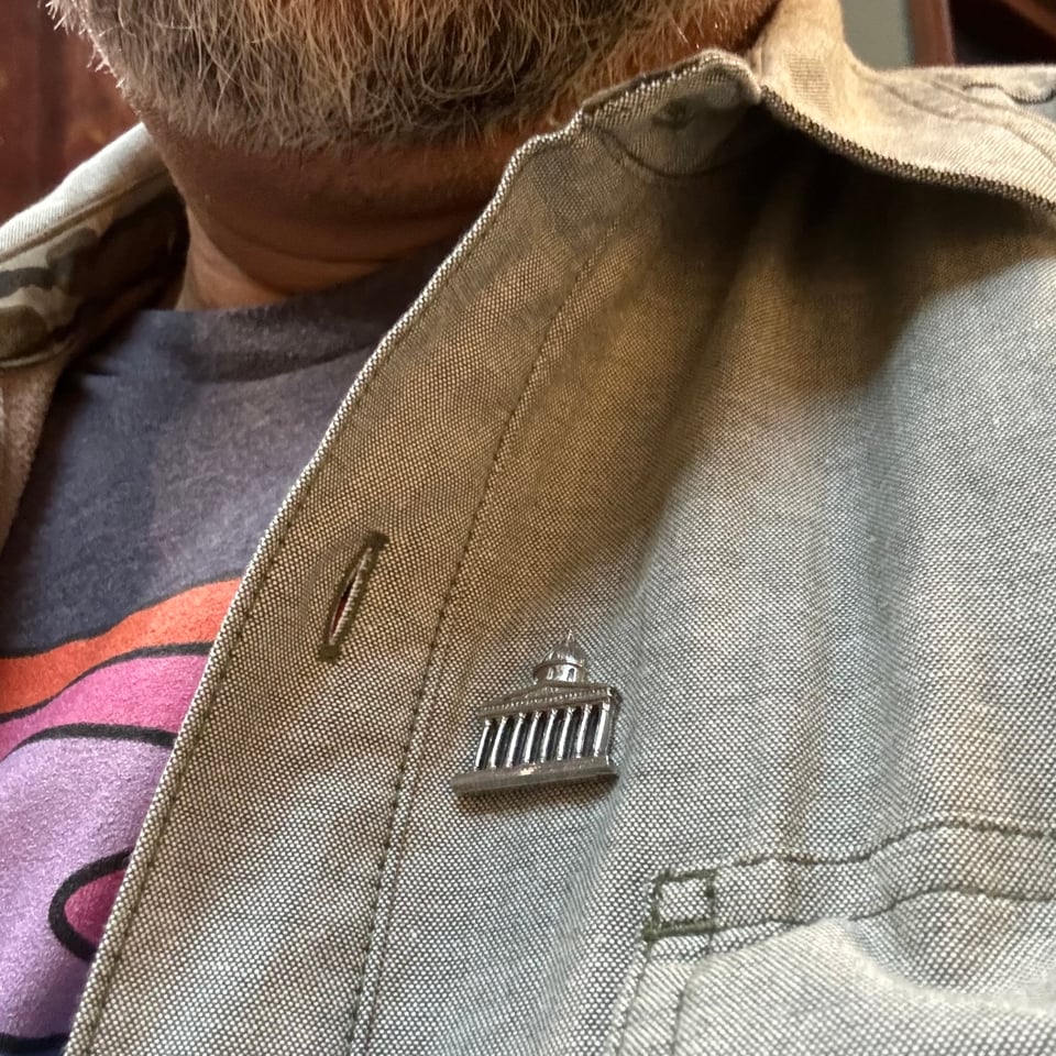 Close-up of pewter pin of the National Gallery facade on an open grey jacket , some of the colourful shirt under the jacket is visible as well as Vinny's neck and greying beard