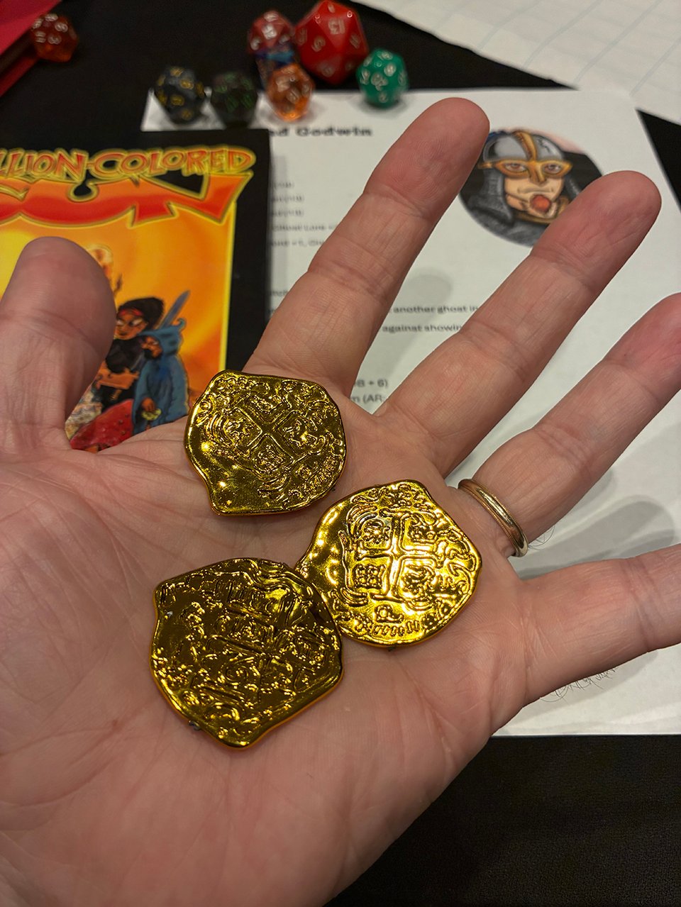Another picture of my hand, for some reason, this time holding some obviously fake gold coins. Weirdly shaped. There's dice on the table, a character sheet is visible, and a copy of Million-Colored Sun. Must be gaming.