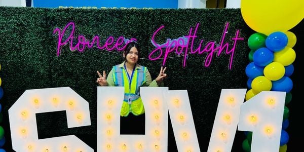 A JMC student wearing a high-visibility safety vest stands behind large illuminated letters spelling 'CVM1'. She is positioned in front of a green hedge wall with pink neon signage reading 'Pioneer Spotlight' and colorful balloons to the side.