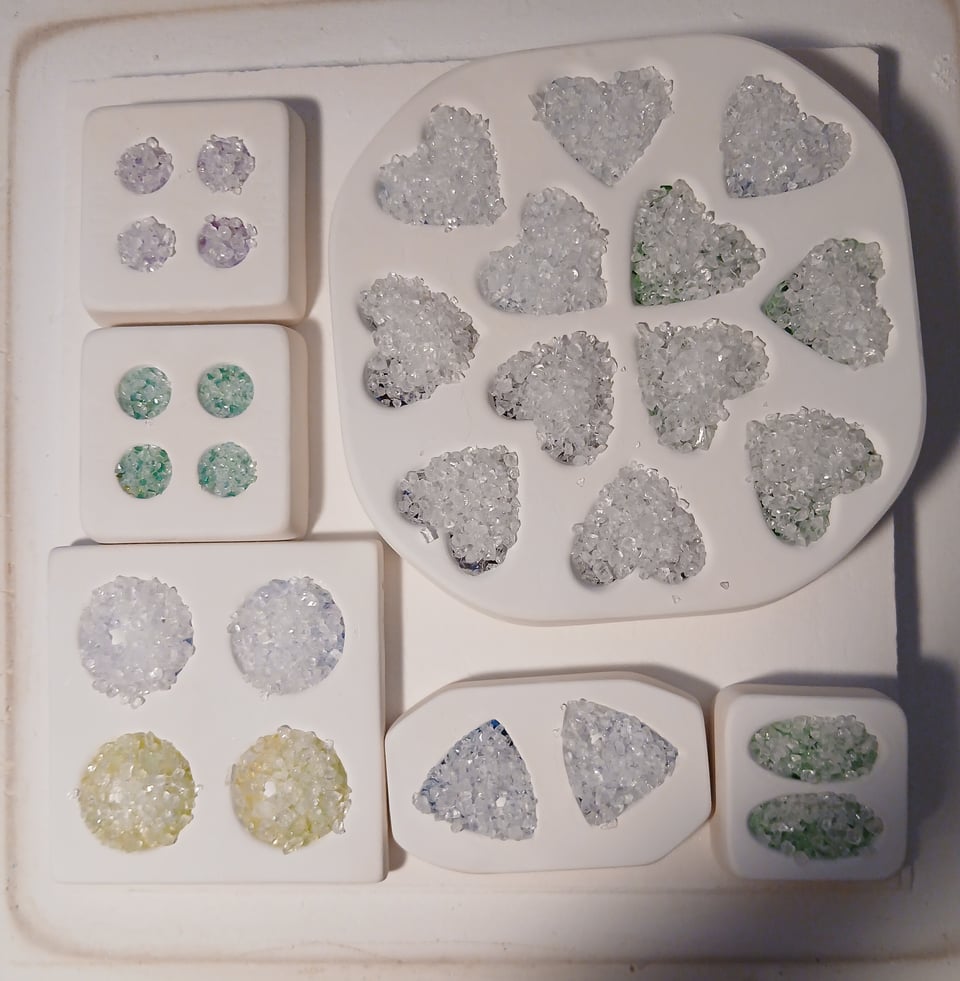 kiln shelf with circle molds in 2 sizes, triangle molds, oval molds and heart molds. most of them have a lot of clear going on but you can see hunts of pink, yellow, blue and green under the clear frit