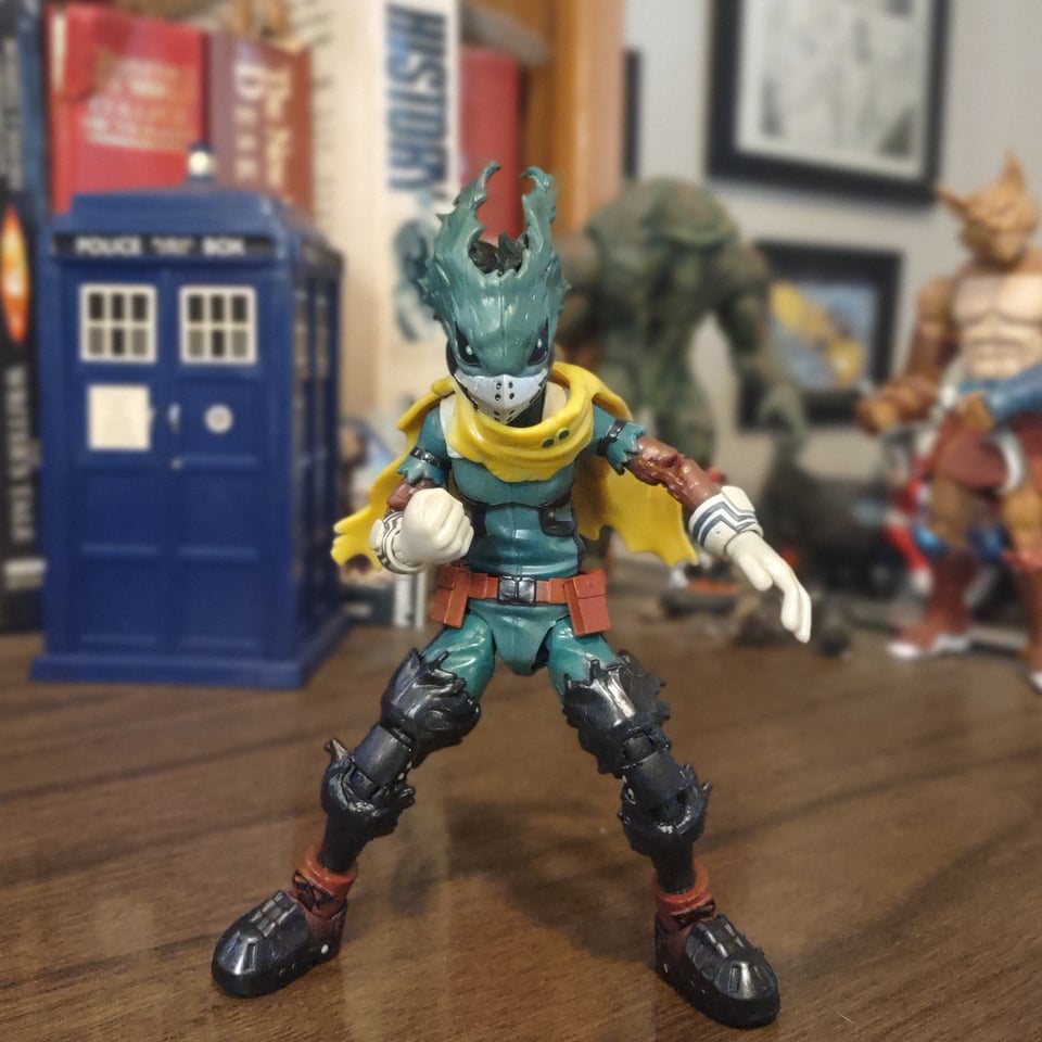 A green and black action figure stands on a wooden desk. It's Dark Deku from the later seasons of My Hero Academia, when he abandons the school and essentially becomes a vigilante. He's wearing a green uniform with thigh-high black boots and white gloves. A yellow scarf, almost a cape, is wrapped around his neck. All of it seems very tattered and uncared for. His face is covered by an equally tattered mask, only showing a muzzle-like mouth guard and two large, dark eyes.