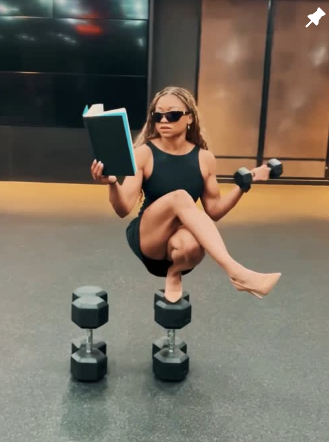 Nadia wearing a sleeveless black sports dress and nude high heels balances with one foot across two dumbbells, with her legs crossed. She holds a book open in one hand and a small dumbbell in the other, looking forward with a serious expression. She wears dark sunglasses, and her hair is styled in long braids. The gym floor beneath her is a speckled black surface, and the background shows dark wall panels with warm lighting.