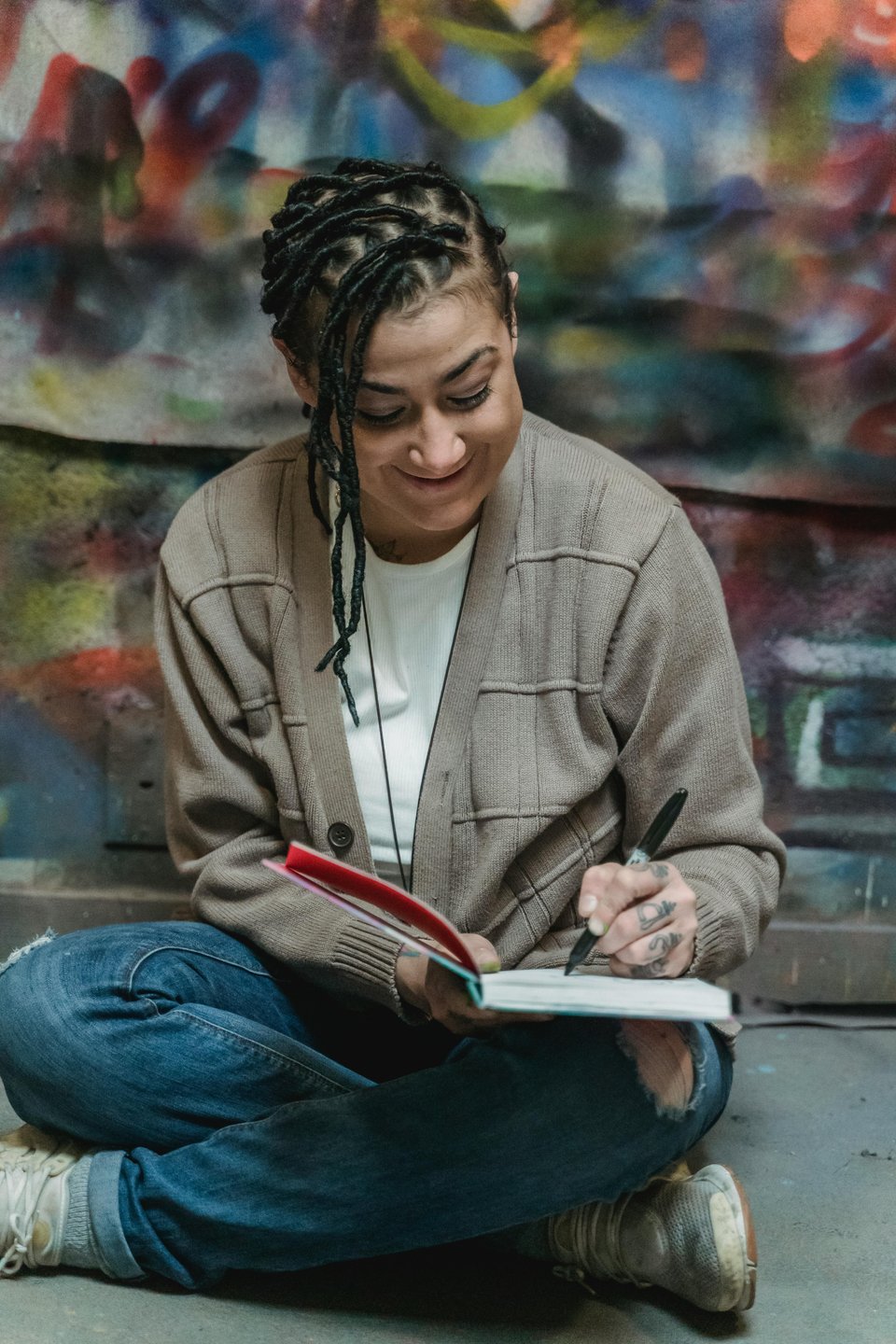 A young woman of color in torn jeans and a beige sweater, smiles as she sits cross-legged as writes with a notebook and pen.