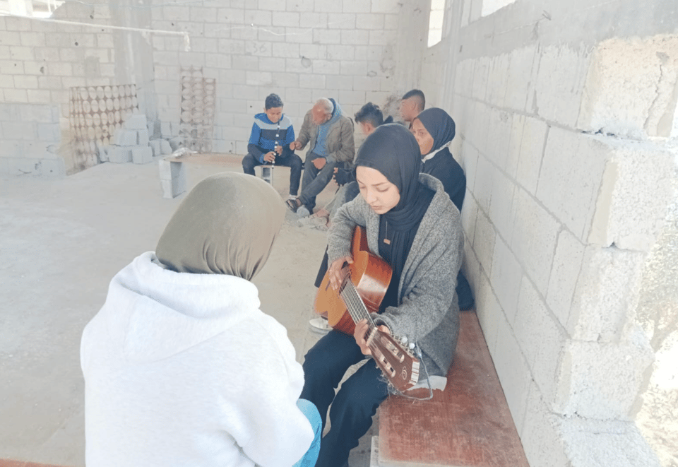 on a bench in a room made of cinderblocks, people seated and two people playing musical instruments 
