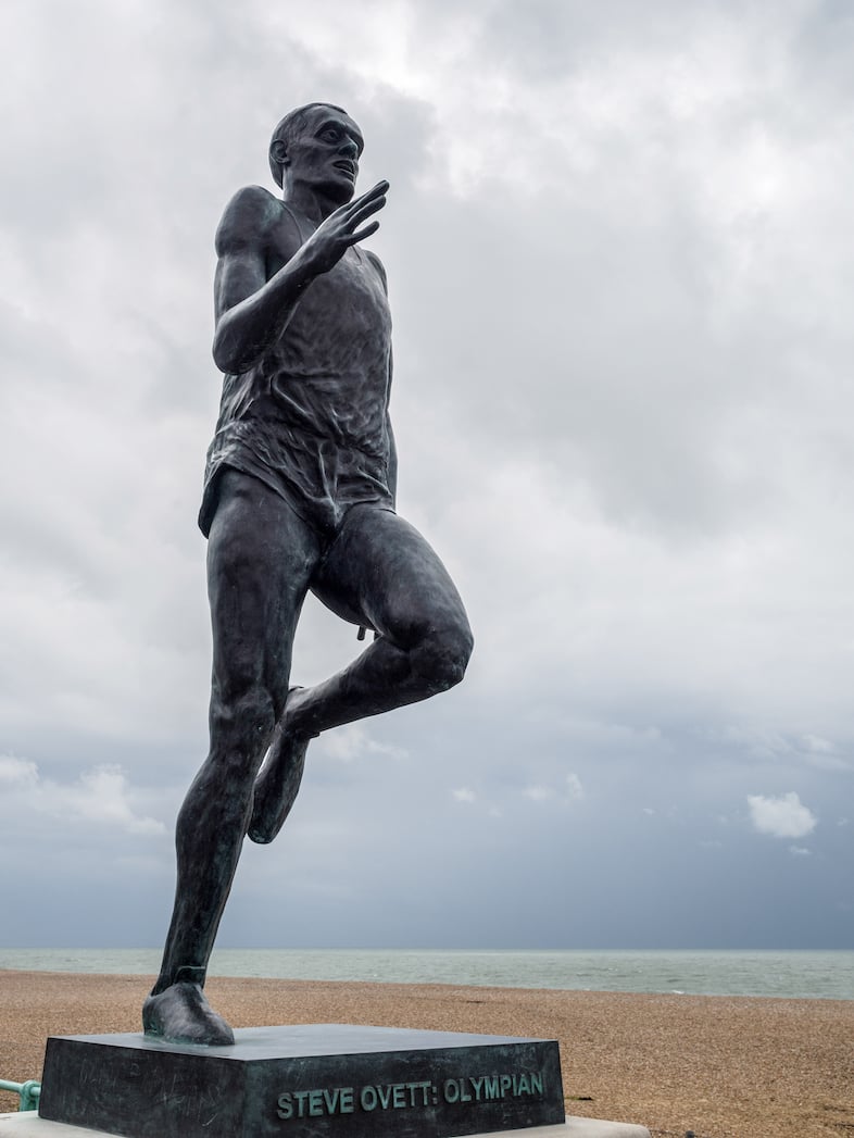 A statue (dull, grey metal) of a runner on a plinth. Text on plinth reads Steve Ovett: Olympian. Beyond is a gravel beach, and then a blue-tree sea.