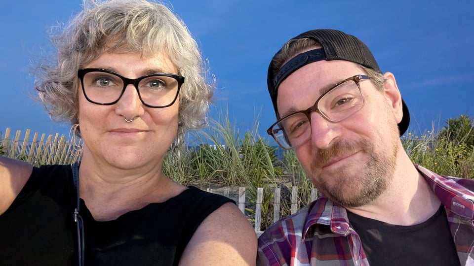 Two white people smile at the camera. One woman has gray hair, glasses and a nose piercing. She's the one holding the camera. The man has a black cap on backwards and glasses. They sit in front of the dune grass at twilight.