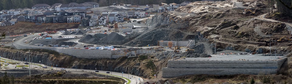 Colour photo of a large construction site. There are no trees, just piles of gravel, dirt, and large retaining walls. There are some houses in the background.