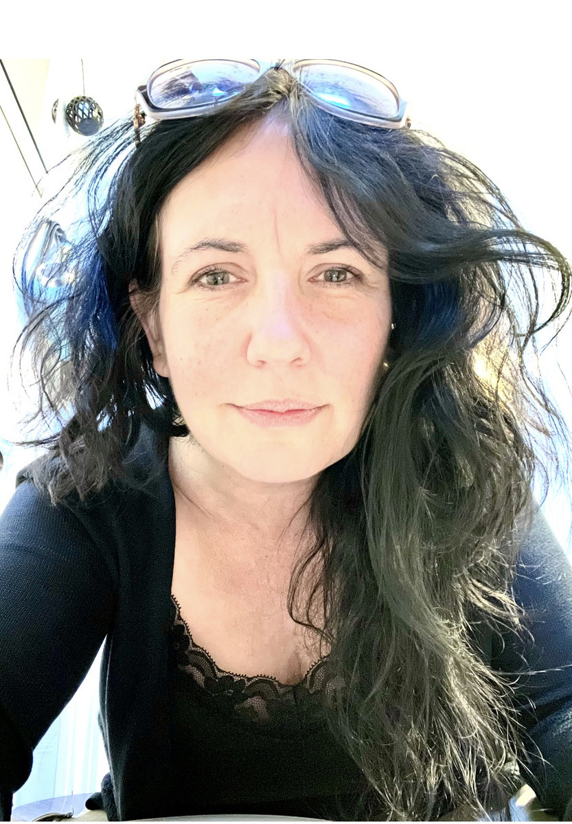 A woman with long, dark hair with sunglasses on top of her head looks directly into the camera.