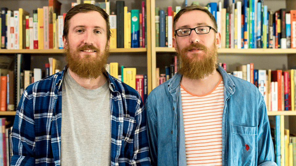 Two men, smiling, side-by-side, looking at the camera. They are identical twins, both with brown straight hair and large bushy beards and mustaches. One is wearing small, rectangular glasses. They stand in front of a bookshelf, filled with books.