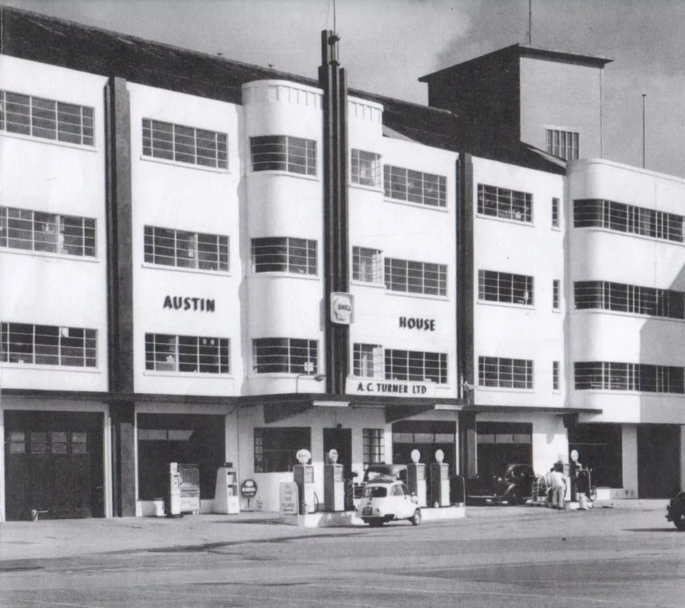 A four storey art deco building. The ground floor is garage bays, with petrol pumps and a bubble car outside. The upper storeys have lots of critall windows, including ribbon ones around the curving bays. A short fin sticks up in the centre.