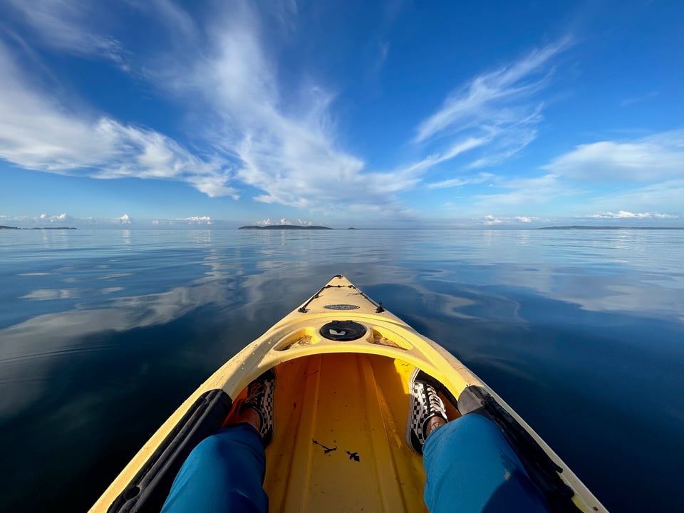 a photo of my legs and the front of a yellow kayak, looking out onto a glassy expanse of ocean with wispy white clouds
