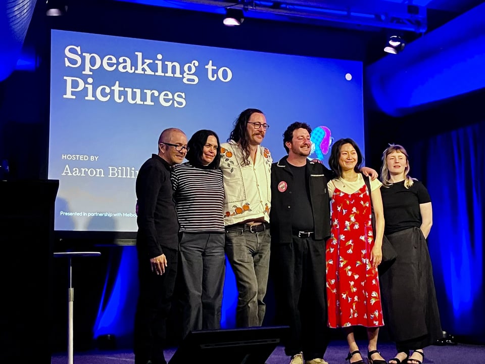 Six people stand shoulder-to-shoulder on a stage, smiling for a group photo in front of a large projected screen reading "Speaking to Pictures — Hosted by Aaron Billings", with blue stage lighting washing the dark walls behind them. The comics creators on stage are: Shaun Tan, Mandy Ord, Joshua Santospirito, Aaron Billings, Rachel Ang, Bailey Sharp