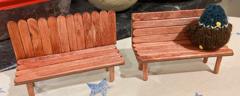 two small park benches made out of popsicle sticks, furniture pegs, and cardboard. On one sits a tiny knitted male mallard duck, very round.