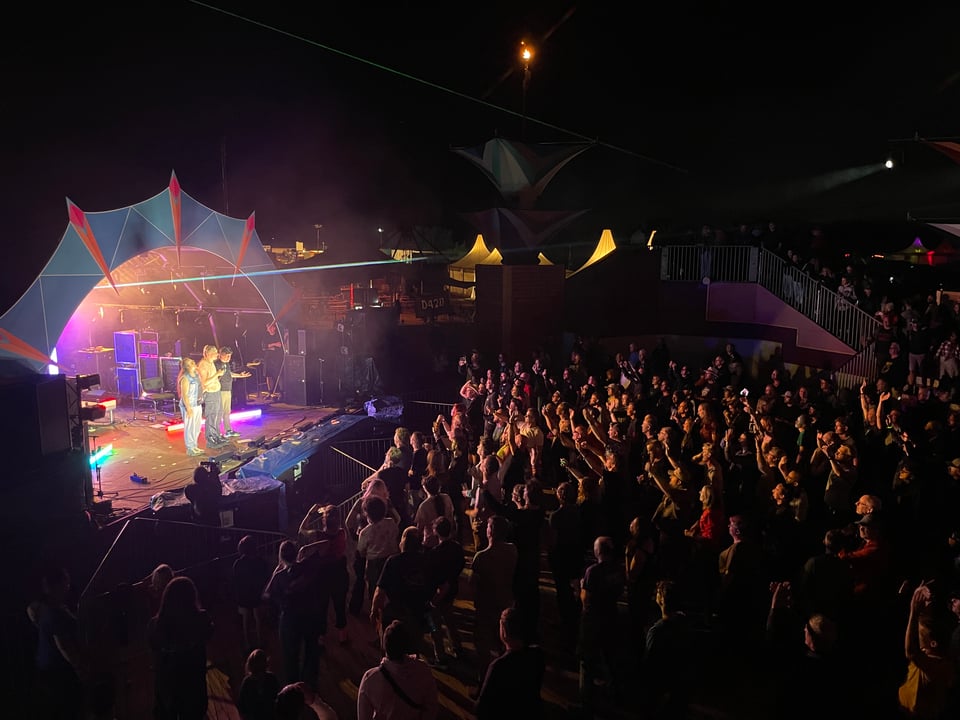 Three musicians on stage thank the audience at the end of their show. The audience's faces are lit up by the stage lights.