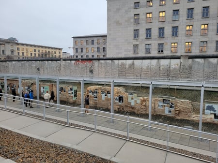 Looking down from an upper level into an exhibition in a trench, tucked in the ruins of a brick building. Above the exhibition is a long piece of the Berlin Wall.