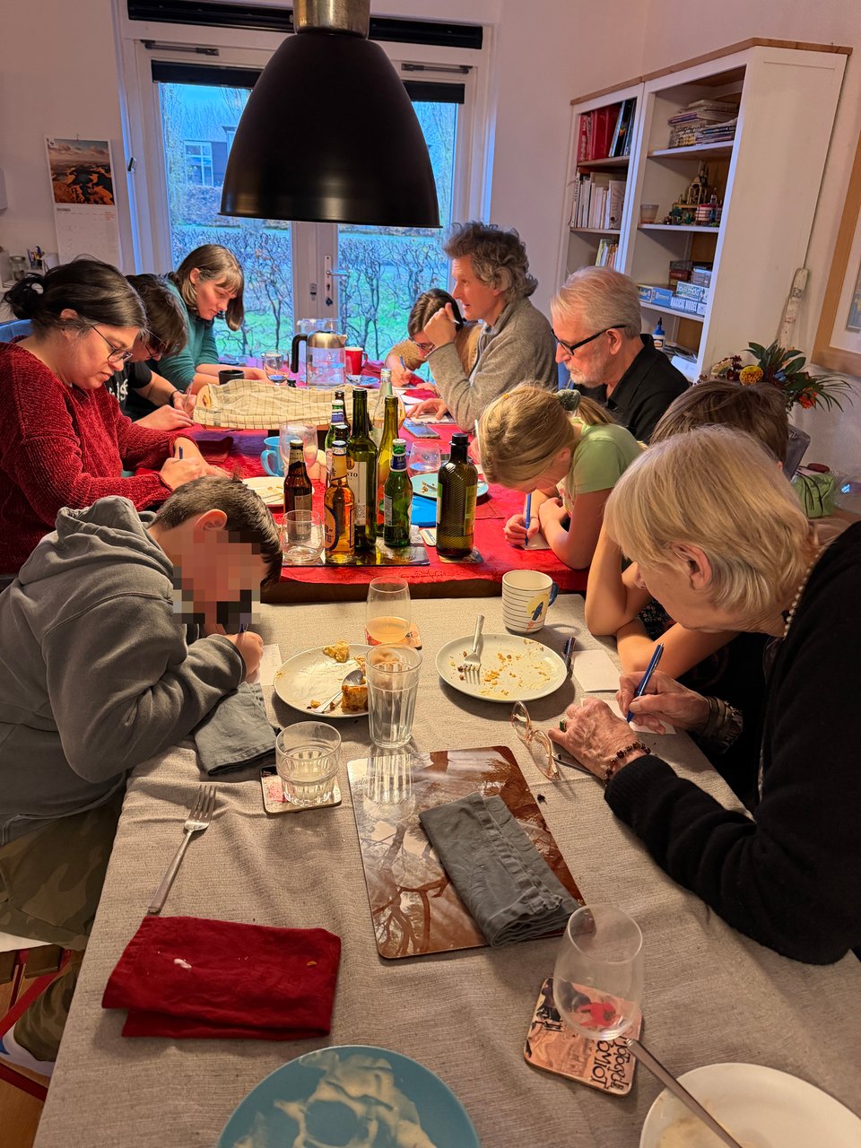 People around a large dining room table are scribbling notes on pieces of paper - playing the game Kim's game trying to remember the items they saw on a tray.
