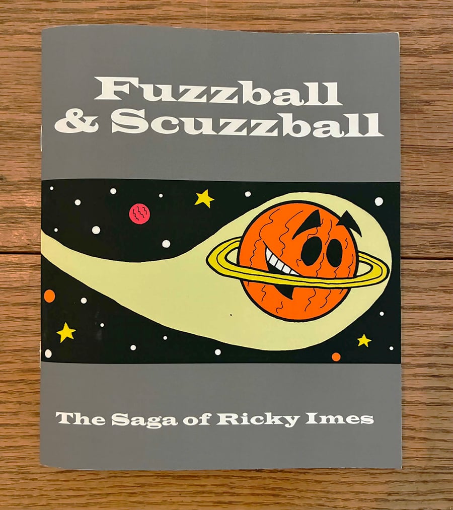 My comic book The Saga of Ricky Imes sitting on a wodden table. The cover is mostly gray, with a picture of Ricky Imes in a band across the middle. Ricky is an orange planet who looks like Saturn with eyes and a mouth, who is soaring through space.